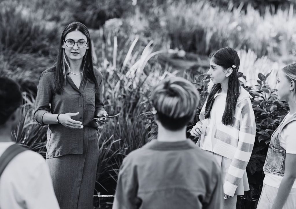 A woman outdoors talking and explaining to a group of youngsters